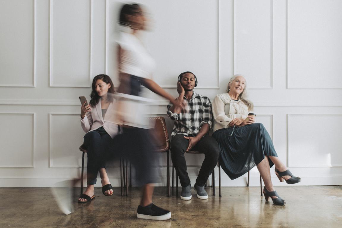In a waiting room, a woman is captured with motion blur as she walks restlessly past other people who are sitting still, illustrating the physical symptom of hyperactivity common with ADHD.