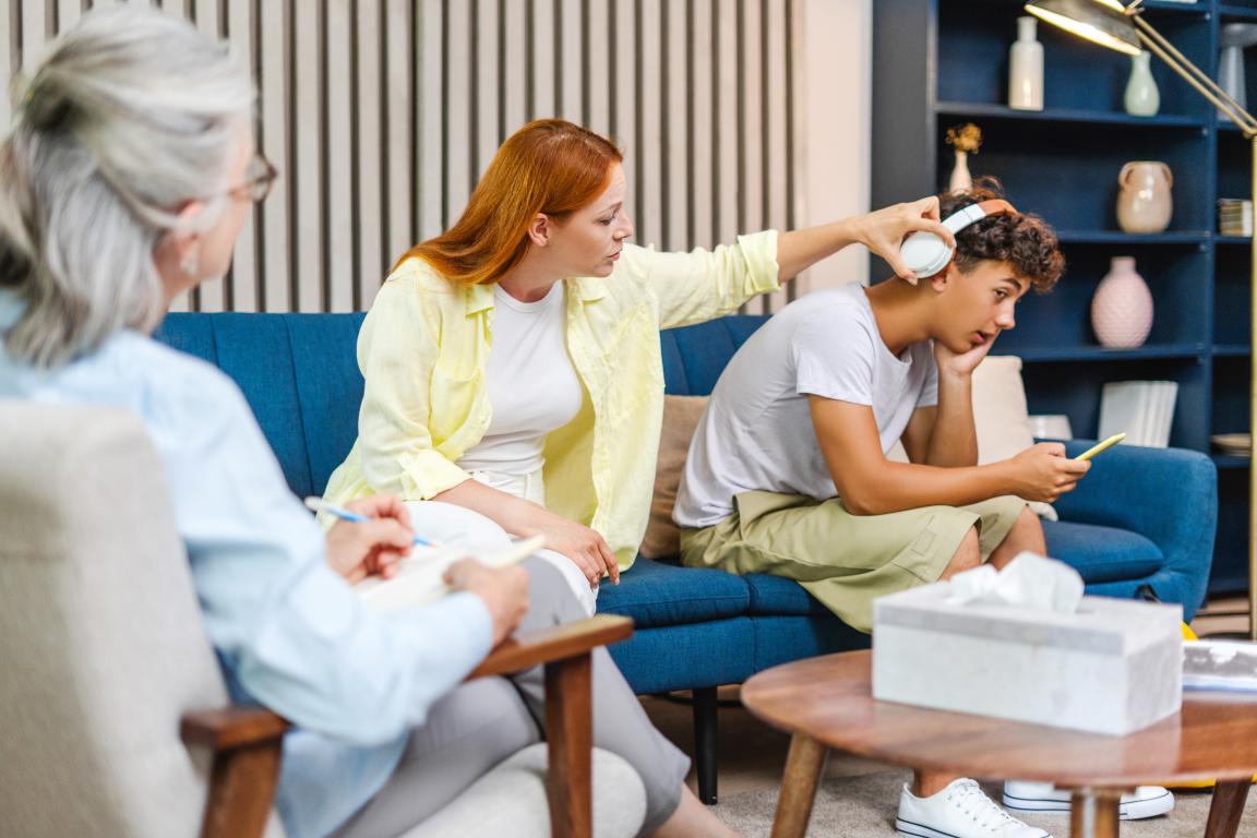 A mom supports her teenage son during a therapy session for ADHD. The son looks withdrawn while his mom gently removes his headphones to help him engage with the female therapist.