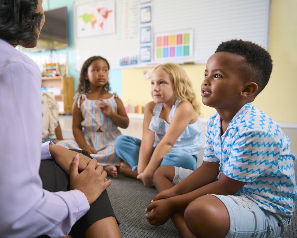 A diverse group of young elementary school children sit on the floor in a circle and listen attentively to their teacher, showing a positive and structured classroom environment beneficial for kids with ADHD.
