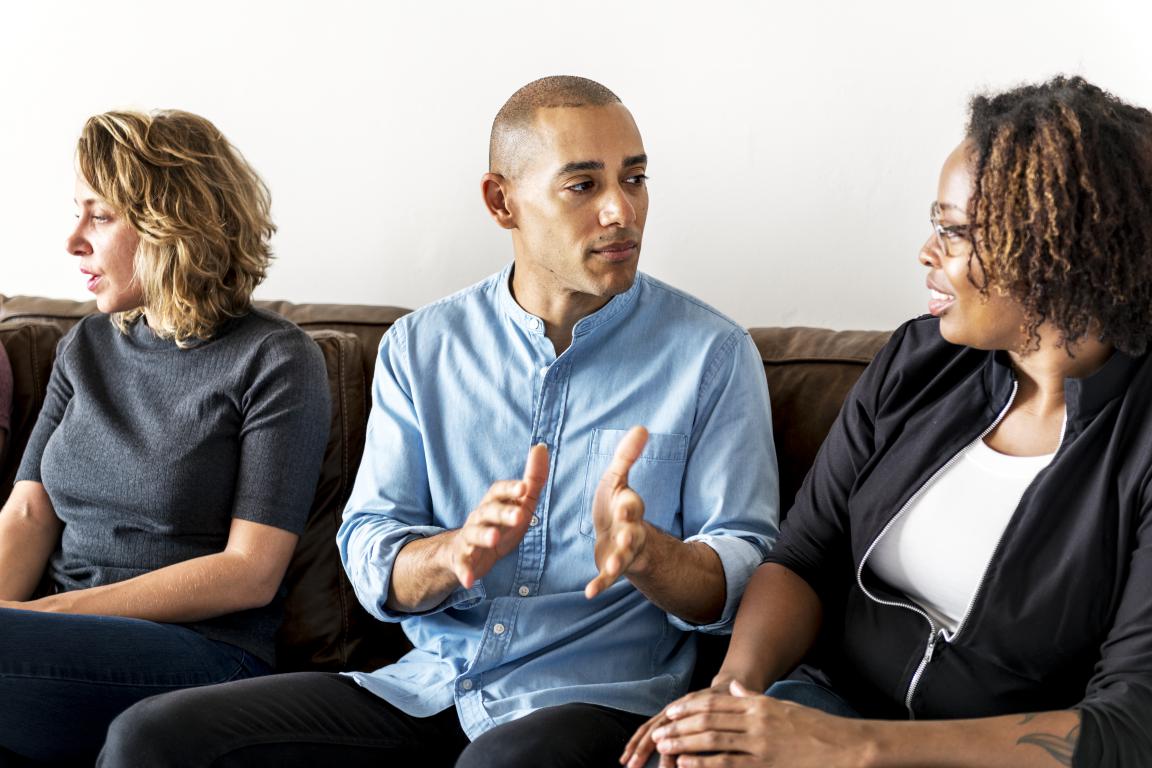 A diverse group of adults sit on a couch during a support group meeting. A man in the center is speaking and gesturing, sharing his experience with others who are listening, highlighting peer support for ADHD.