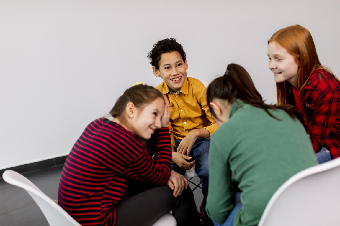 A diverse group of pre-teens laugh and talk while sitting in a circle of chairs during a peer support group, demonstrating the benefits of shared experiences for children with ADHD.