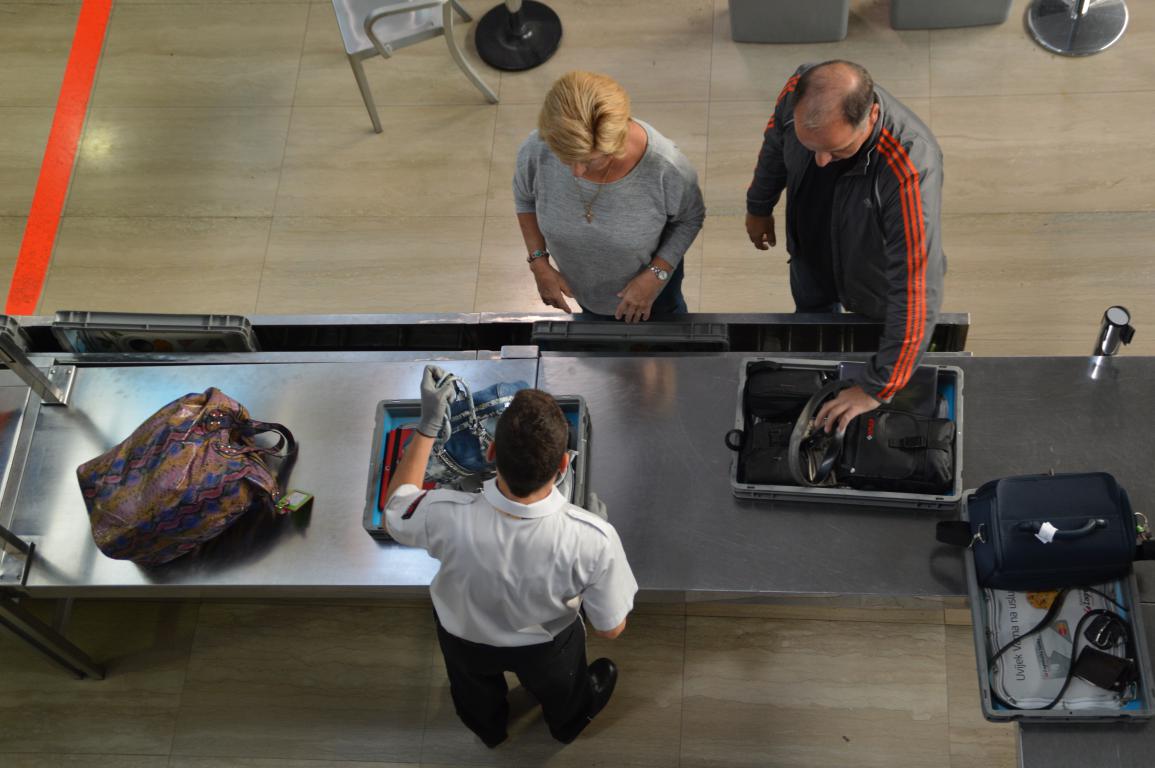 An overhead view of a couple at an airport security checkpoint, placing their bags and belongings into trays on the conveyor belt as a security officer assists them.