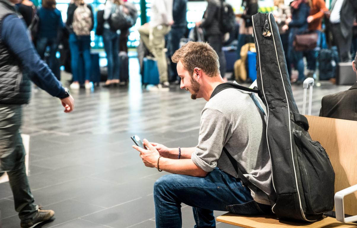 A man with a guitar case on his back sits calmly on a bench in a busy airport departure lounge, smiling as he looks at his phone. This represents finding a calm moment while waiting for a flight.