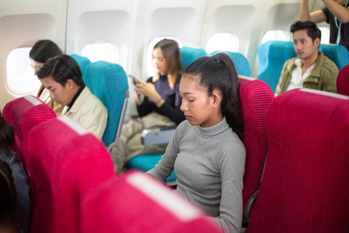  A young woman with her eyes closed rests in her seat on a crowded aeroplane, managing the stress and sensory input of being on a flight. Other passengers are blurred in the background.