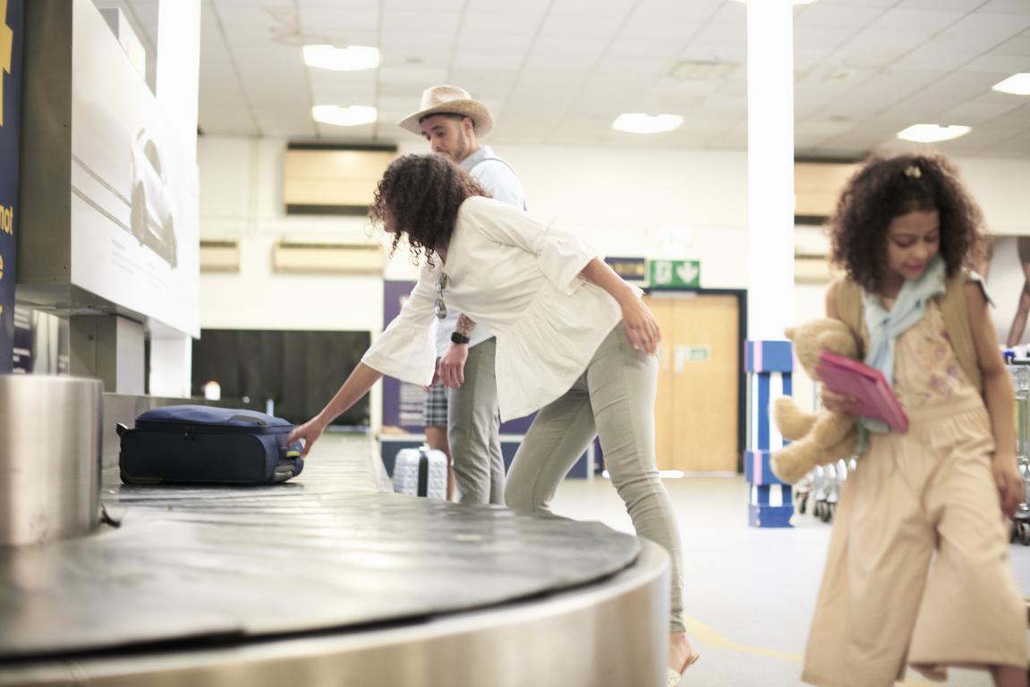 A family with a young daughter waits at the baggage reclaim carousel in an airport arrivals hall. The mother is reaching for their suitcase as it comes around, demonstrating the final stage of the journey.