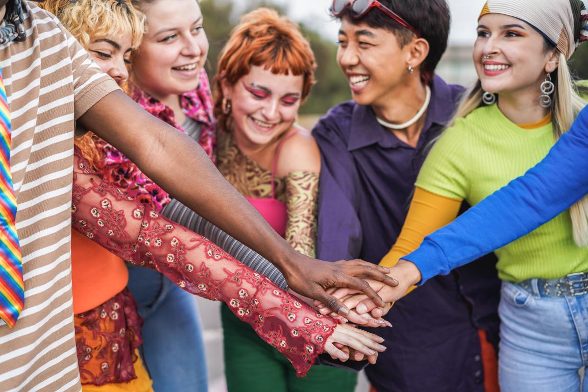 A diverse group of young people stand in a circle with their hands together in the middle, smiling and showing solidarity. This represents finding community, support, and helpful resources for ADHD.