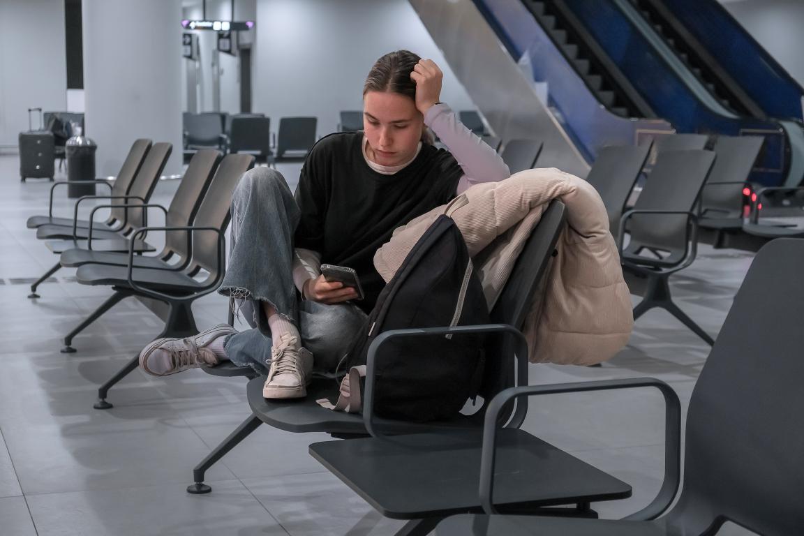 A person sits slumped in a chair in an empty airport departure lounge, looking down at their phone with a stressed or tired expression. This depicts the feeling of anxiety that can occur while waiting for a flight.