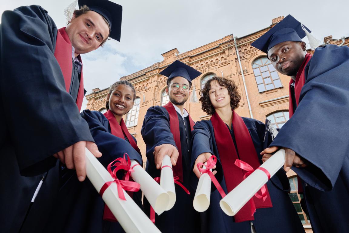 A group of diverse university graduates stand together smiling holding their diplomas in front of a building