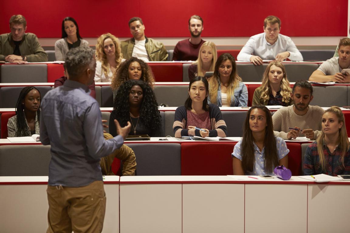 A male professor stands at the front of a lecture hall, gesturing while teaching a group of diverse university students