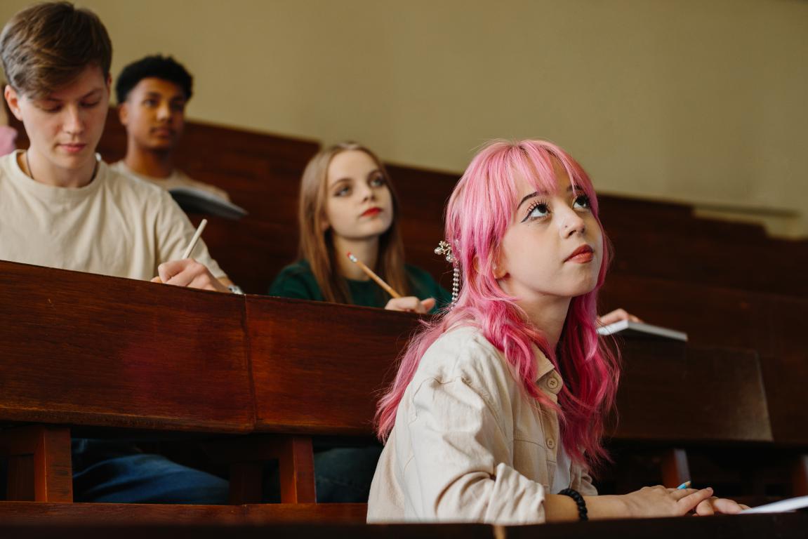 A young woman with pink hair sits in a classroom looking upwards with other students around her