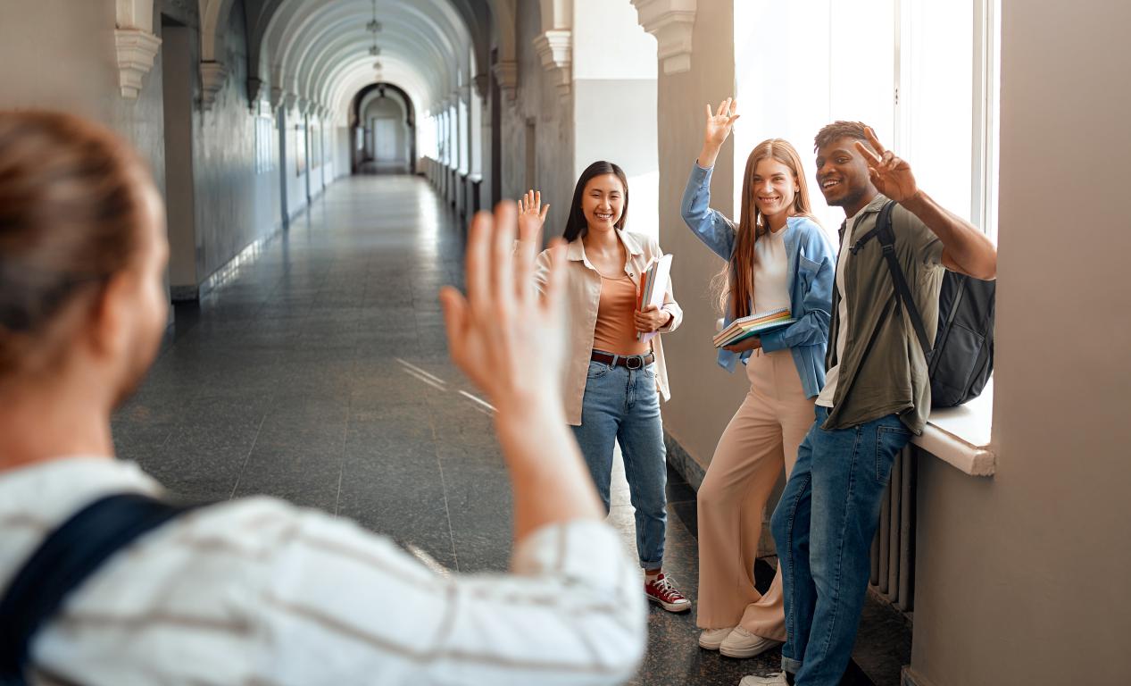A group of four university students are in a hallway with one student waving to someone