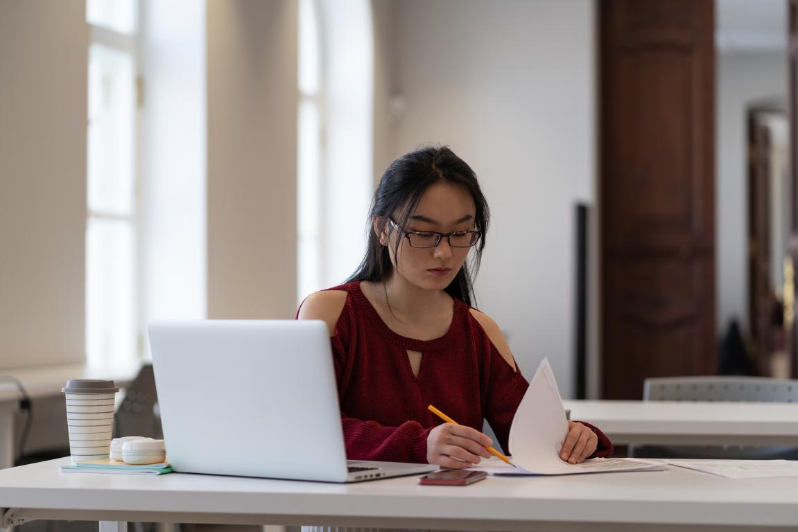 A young woman with glasses sits at a table and writes in a notebook, with a red cup nearby