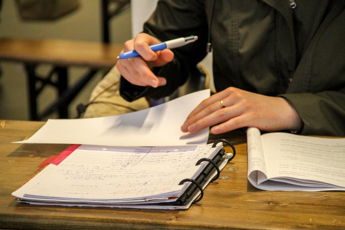 A person's hands are shown writing in a spiral-bound notebook with a pen, with other books and papers on the table
