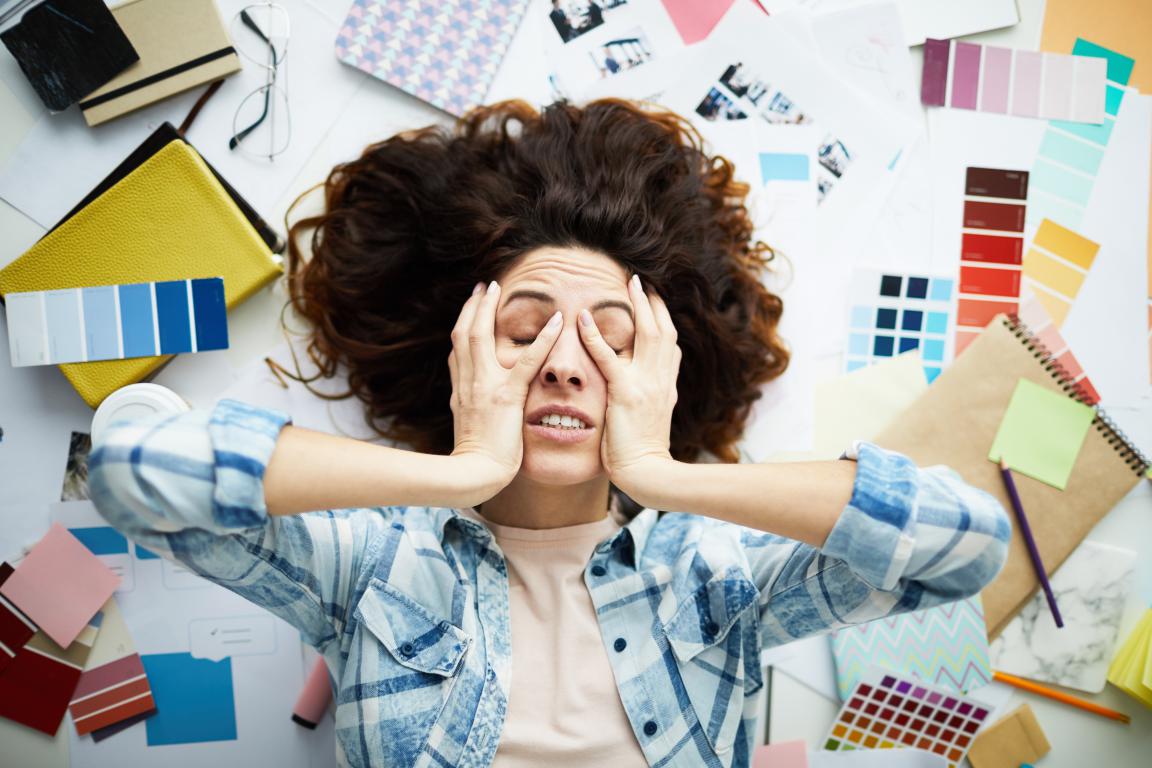 A woman lies on the floor surrounded by a chaotic mess of papers and creative supplies, her hands on her face in an expression of complete burnout and overwhelm.