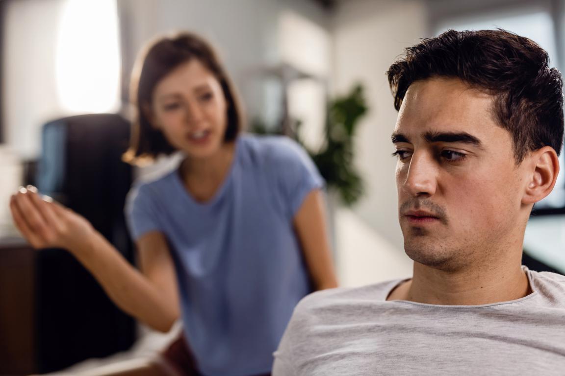 A young man looks away with a hurt and withdrawn expression while a woman in the background speaks to him with frustration.