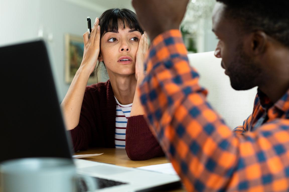 A woman looks up with a worried and overwhelmed expression, hands on her head, as a man in a plaid shirt speaks to her animatedly across a desk