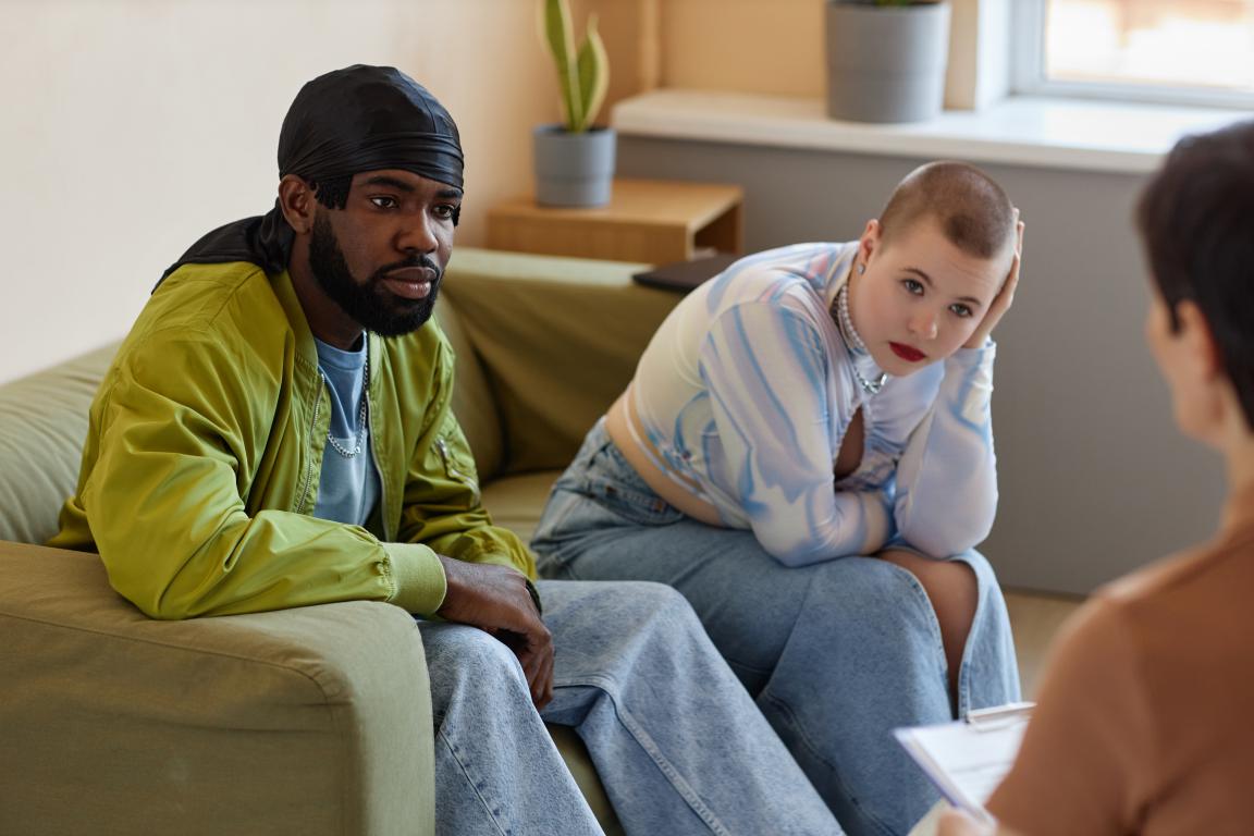 A young man and woman sit on a sofa during what appears to be a therapy session, both with serious and contemplative expressions.