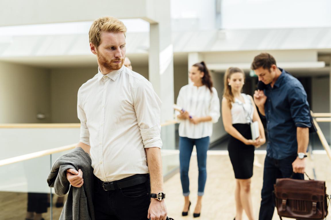 A man looks back with a pained expression at a group of people talking without him in an office hallway, capturing a sense of being excluded