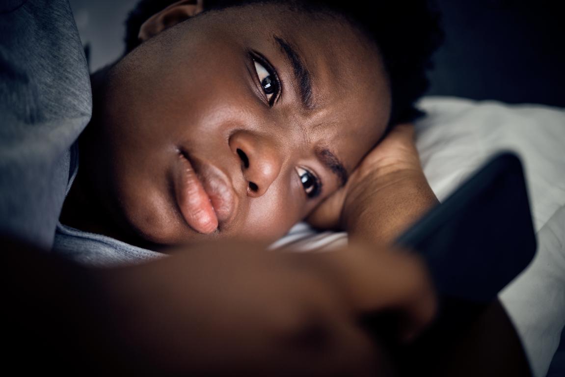 A close-up shot of a young Black person lying in bed, their face illuminated by their phone, with a worried and furrowed brow as they stare at the screen.