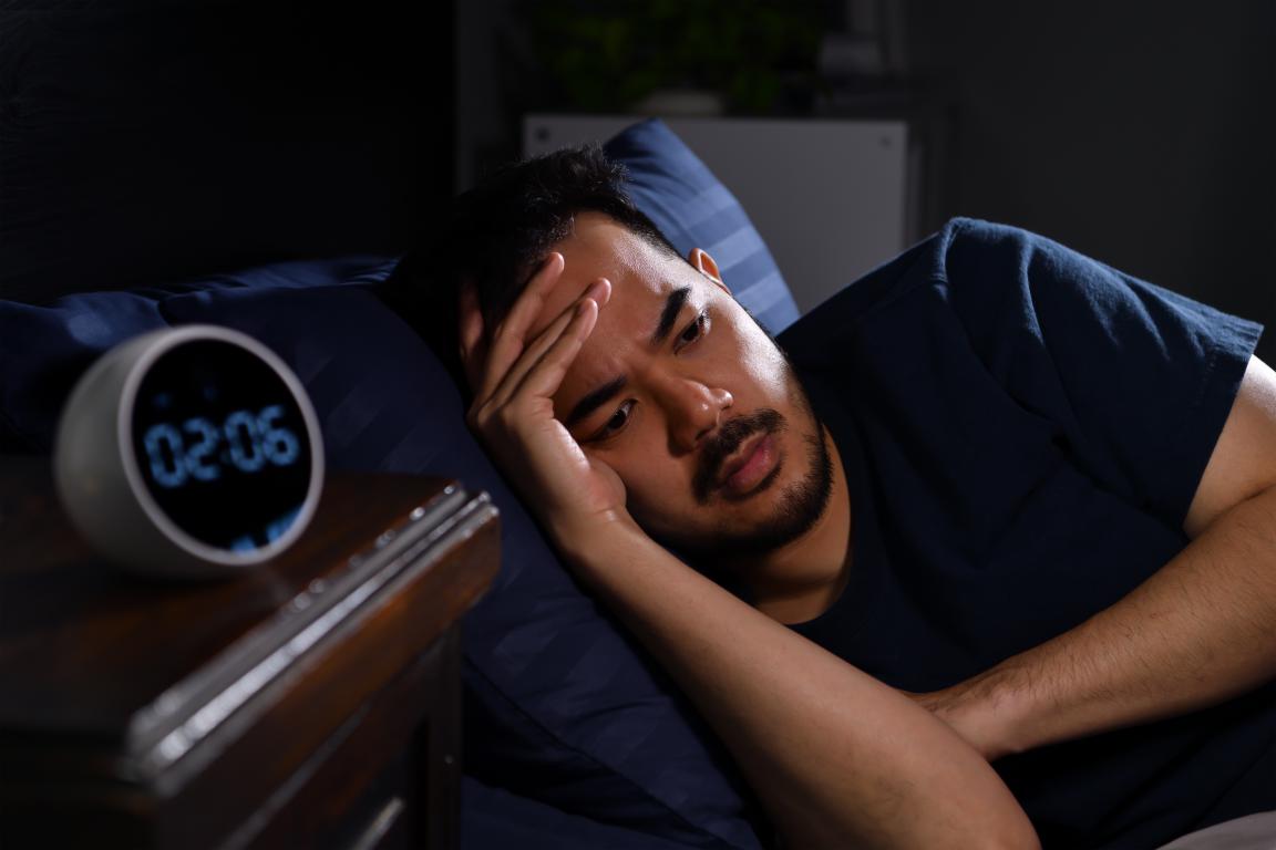 An anxious man lies awake in a dark room, his hand on his forehead, looking at a digital clock on his bedside table that reads 02:06 AM.