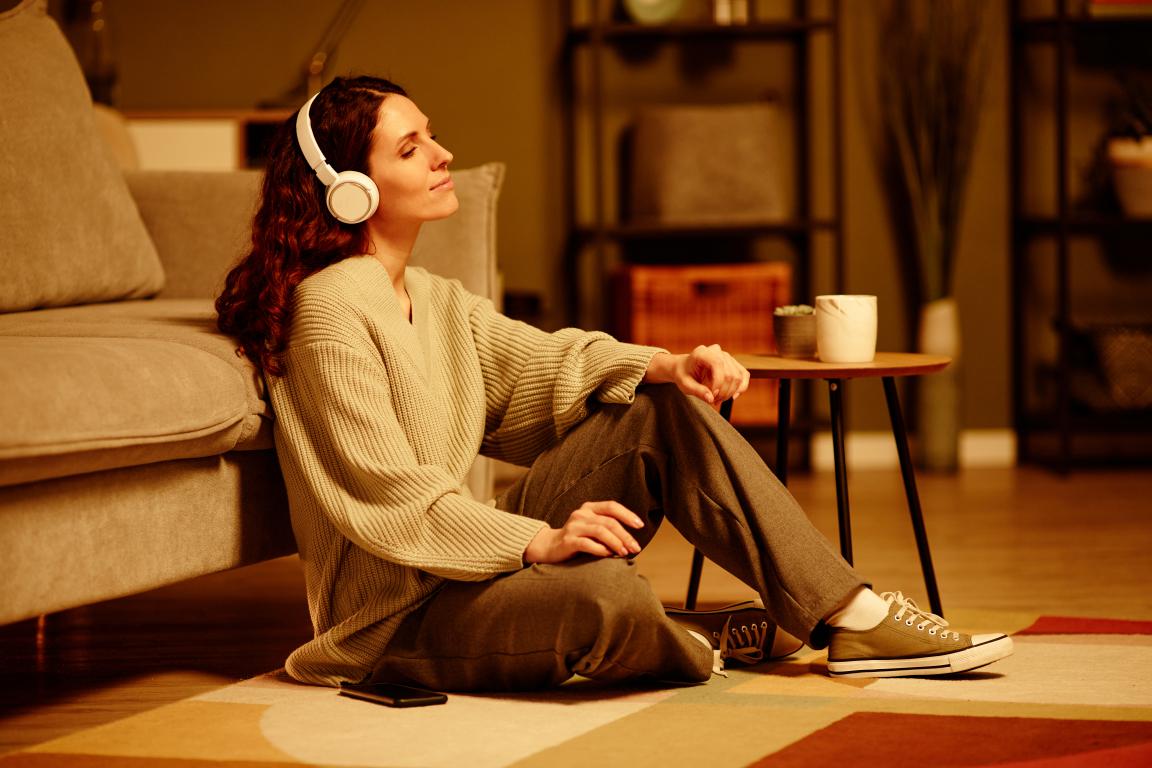 A relaxed woman with her eyes closed sits on the floor of her cosy living room, peacefully listening to something on her headphones as part of a wind-down routine.