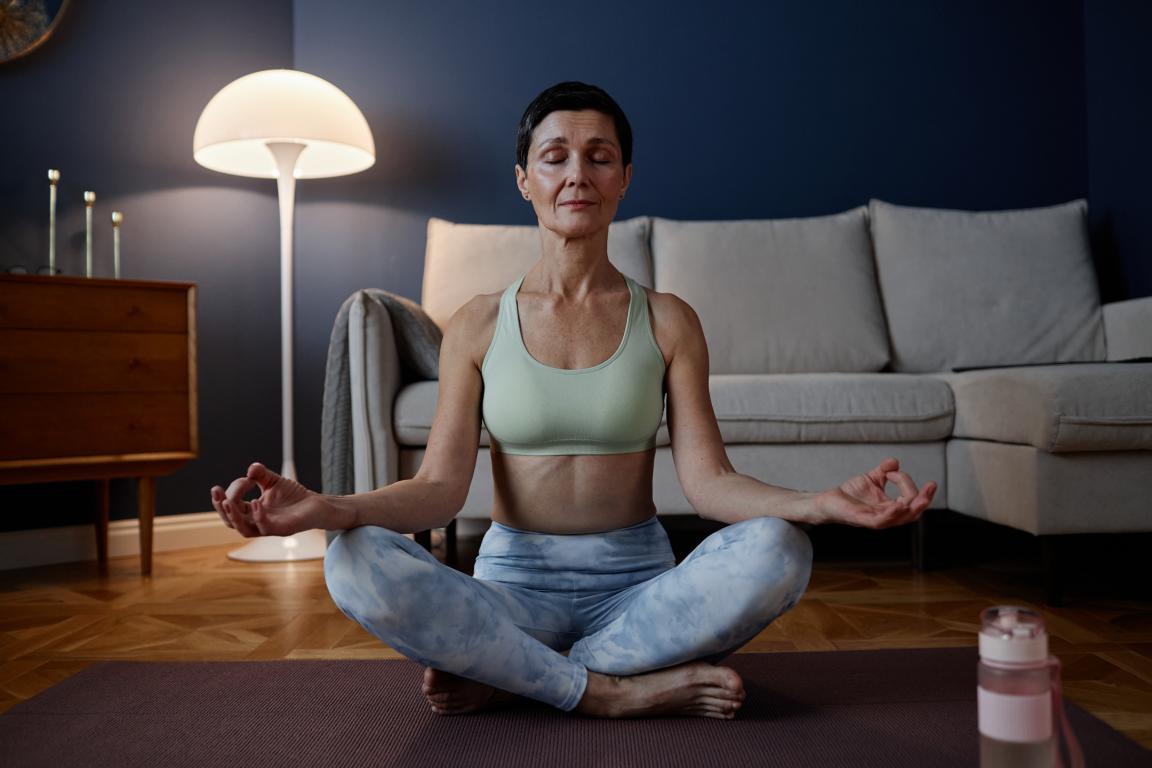 A calm woman with short hair meditates in a lotus position on a yoga mat in her living room, with her eyes closed and a peaceful expression.