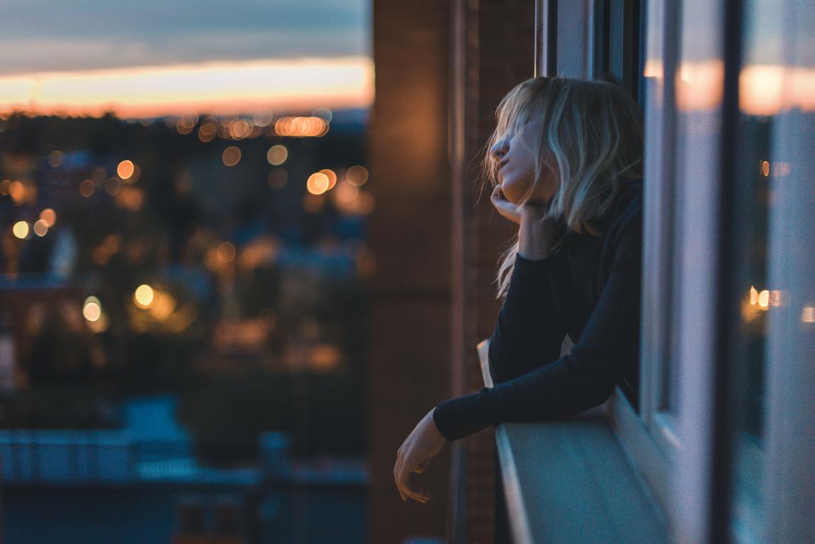 A woman with blonde hair leans out of a window at dusk, looking thoughtfully over a city skyline with blurred lights in the background.