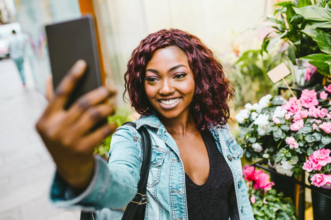 A smiling Black woman with curly reddish-purple hair takes a selfie outdoors in front of a vibrant display of pink and white flowers