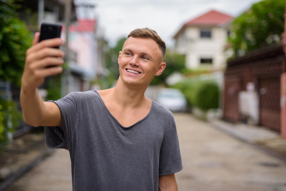 A smiling young man in a casual grey t-shirt stands on a residential street and takes a selfie with his smartphone.
