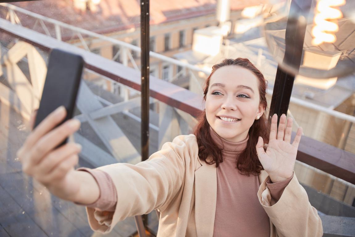 A young woman with reddish-brown hair smiles and waves at her phone as she takes a selfie from a high vantage point, with a city visible below.