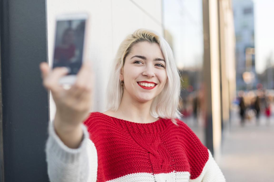 A woman with short platinum blonde hair and red lipstick smiles while taking a selfie on a city street next to a modern building.