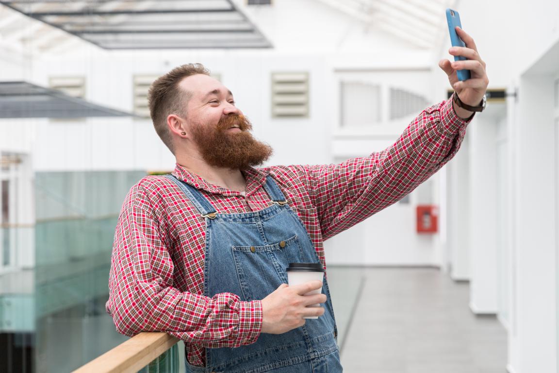 A smiling man with a large red beard, wearing denim dungarees and a plaid shirt, takes a selfie in a bright, modern office hallway.