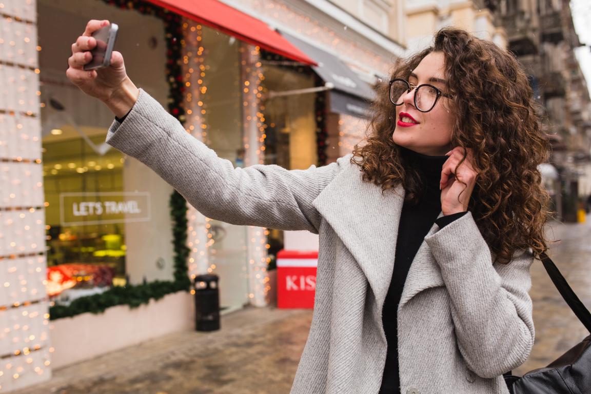 A stylish woman with curly hair and glasses smiles as she takes a selfie on a city street with lit-up shop windows in the background.