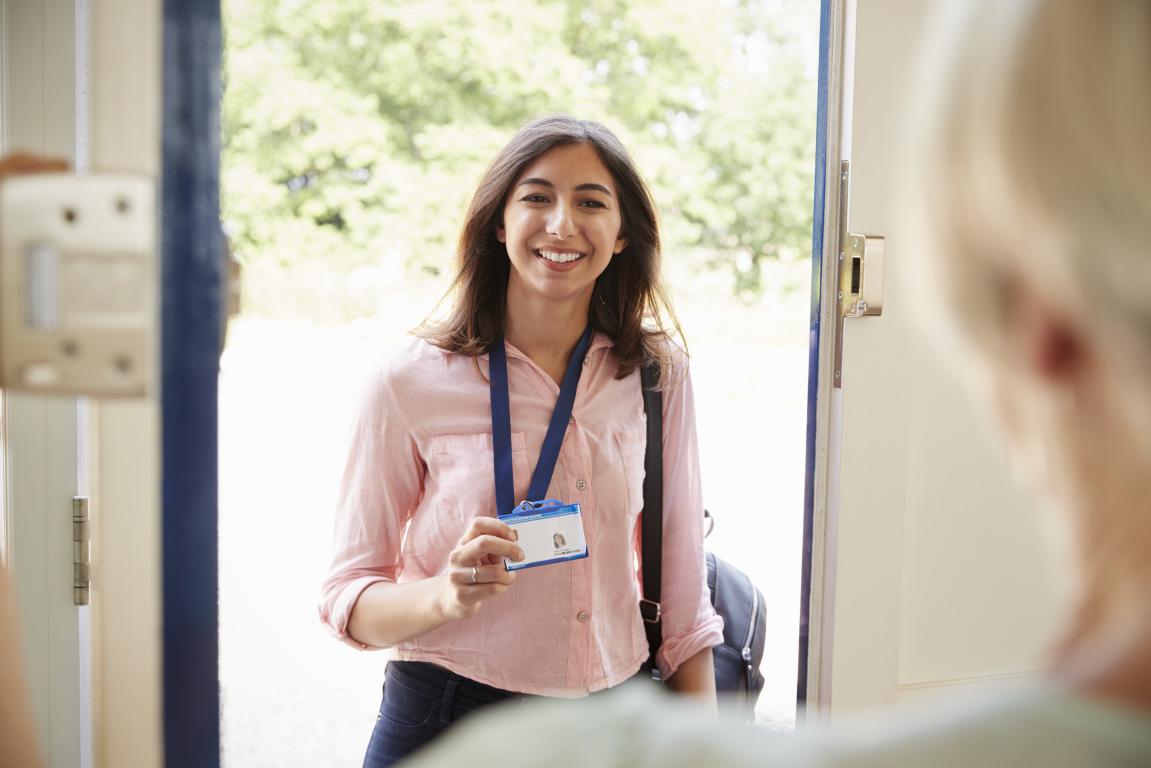 A friendly support worker wearing a professional ID card on a lanyard smiles as she is welcomed into a client's home.