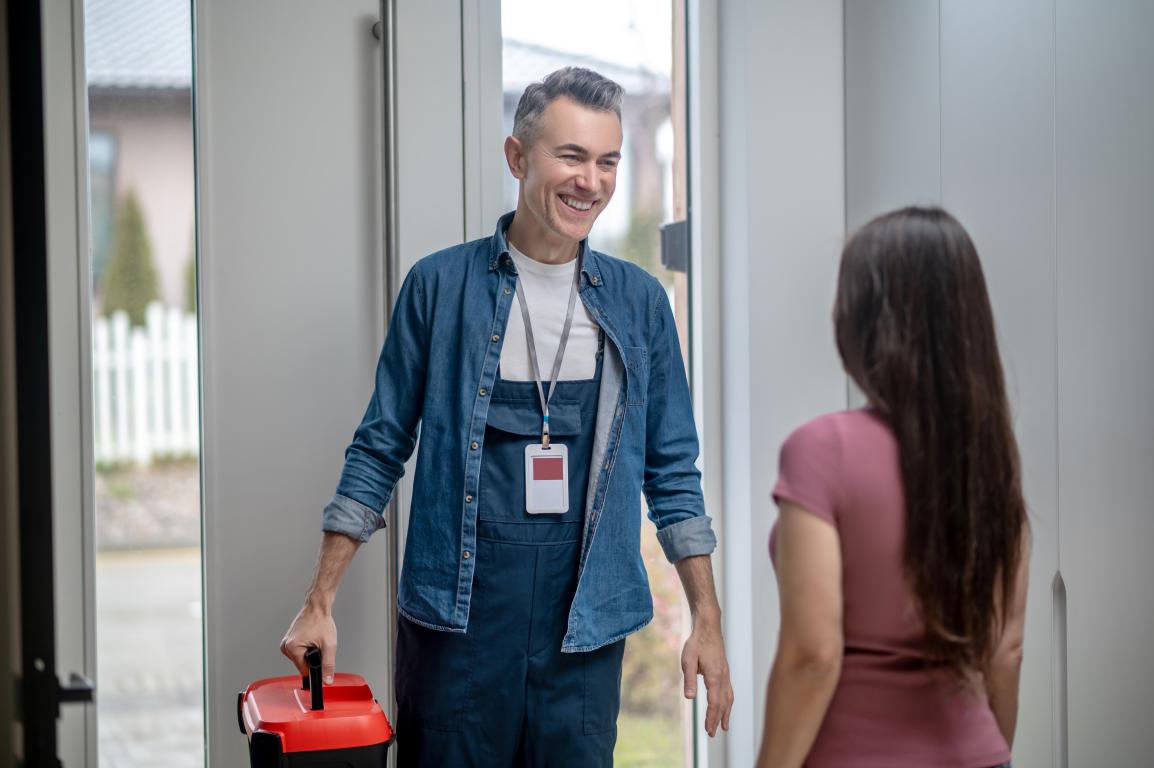 A professional technician wearing an ID card and carrying a toolbox smiles at a client in her home, ready to start the job.