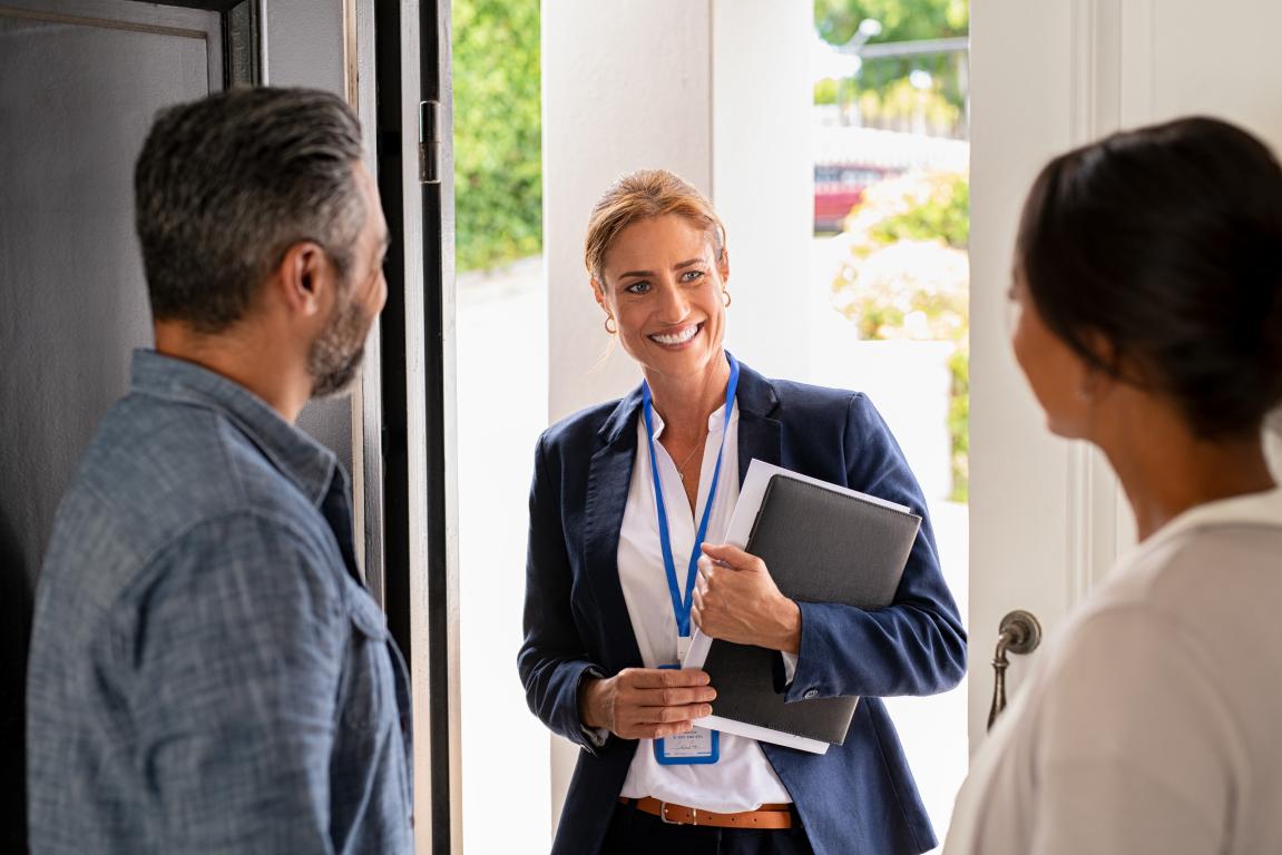  A female business professional wearing a blazer and an ID card on a lanyard greets a couple at the door of their home.