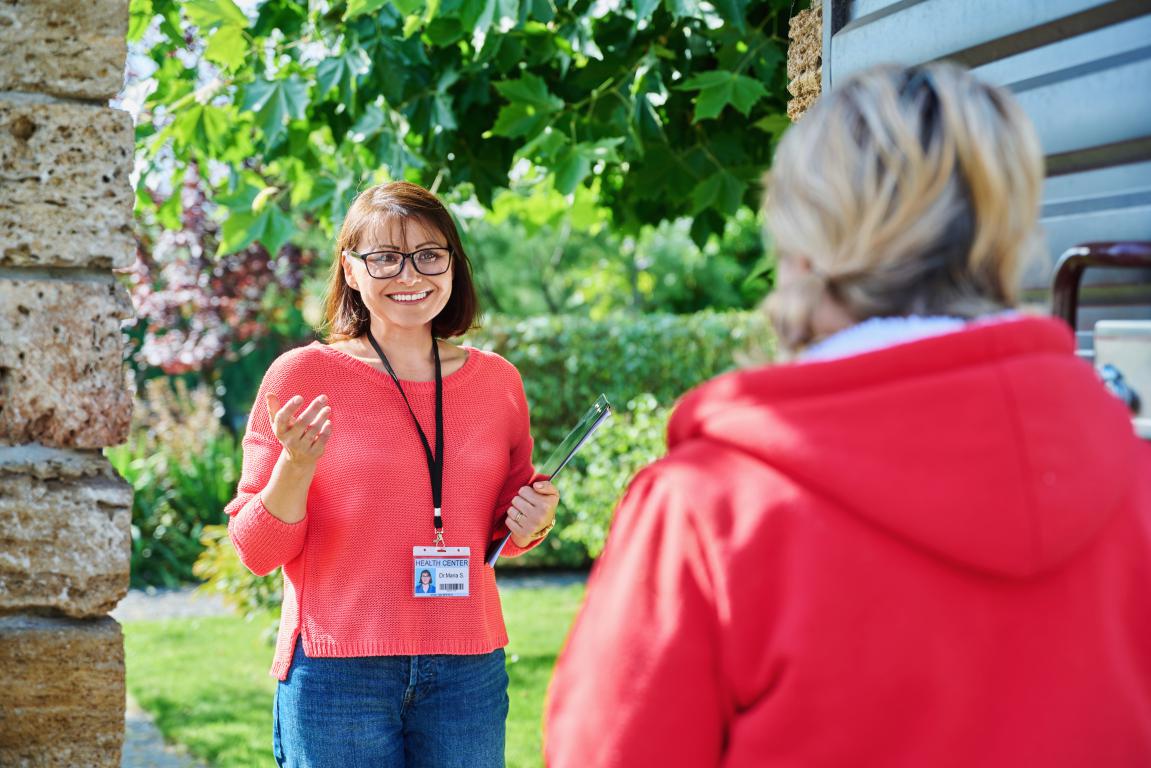 A health center employee with an ID card and clipboard speaks with a client at her front door during a home visit.
