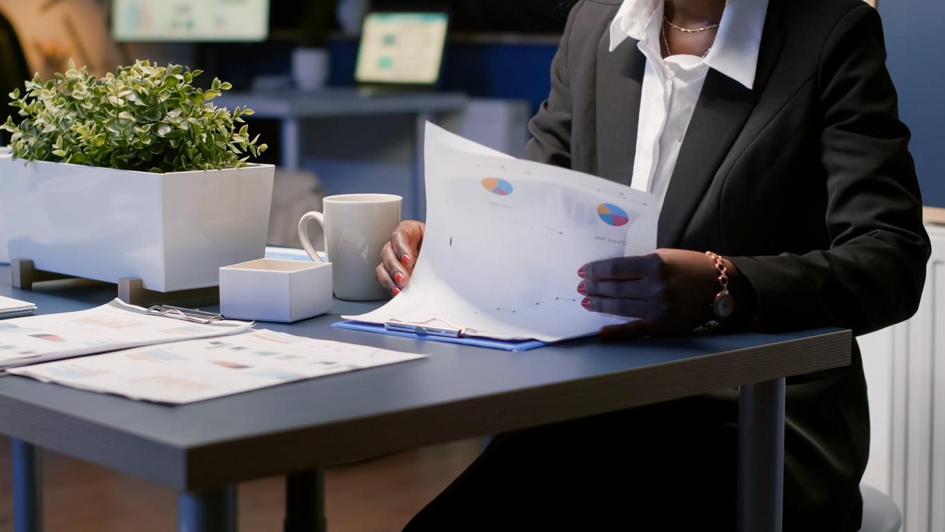  A professional woman in a business suit sits at her desk in an office, carefully reviewing documents with colorful pie charts and graphs.