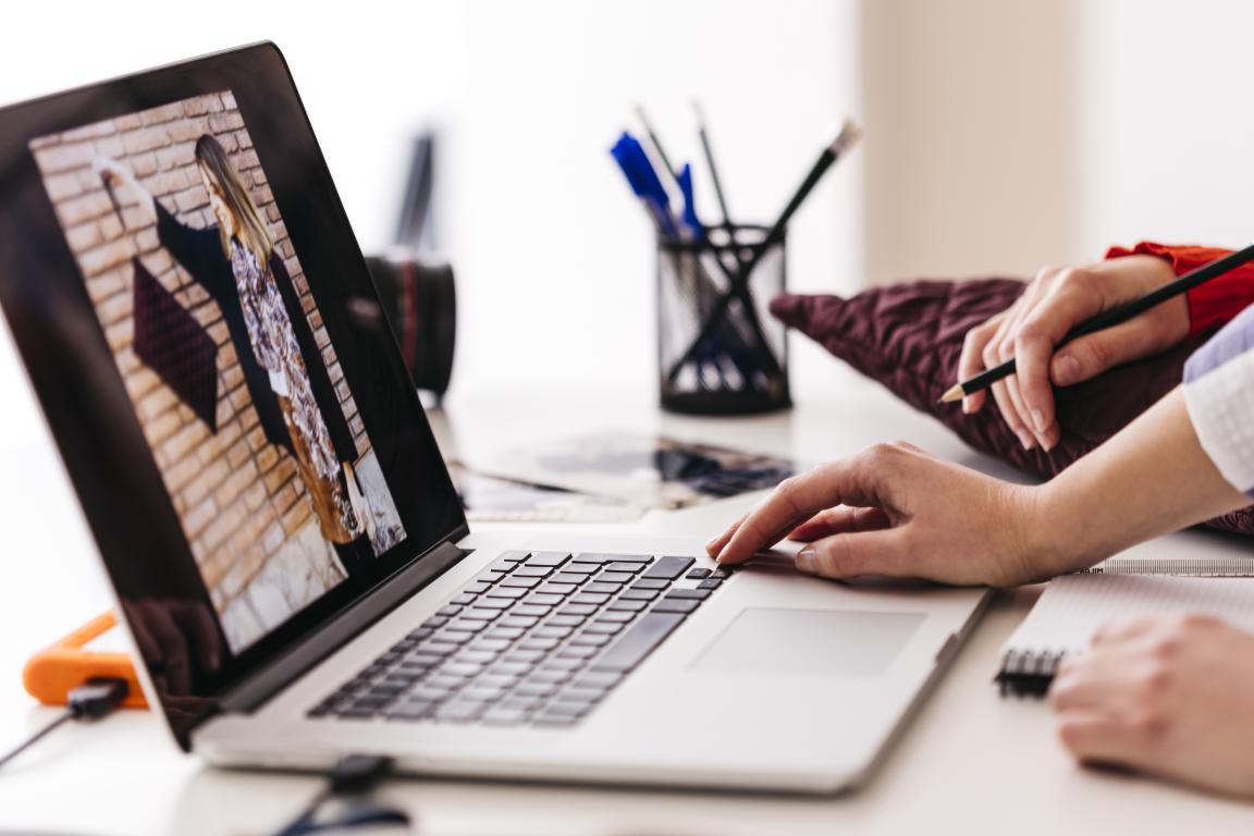 A person's hands work on a laptop, editing a fashion photo, with a pencil and notepad nearby suggesting a creative design process.