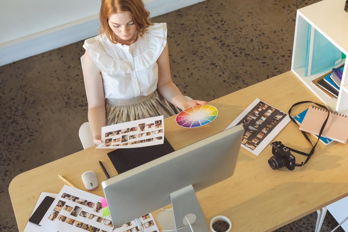 From an overhead view, a female graphic designer sits at her desk, comparing a sheet of photos with a color wheel to make branding decisions.
