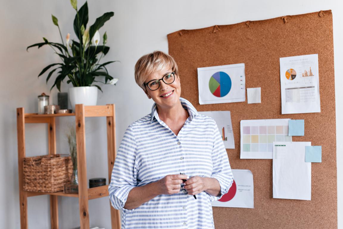 A confident female professional with glasses smiles while standing in front of a corkboard displaying business charts in her office.