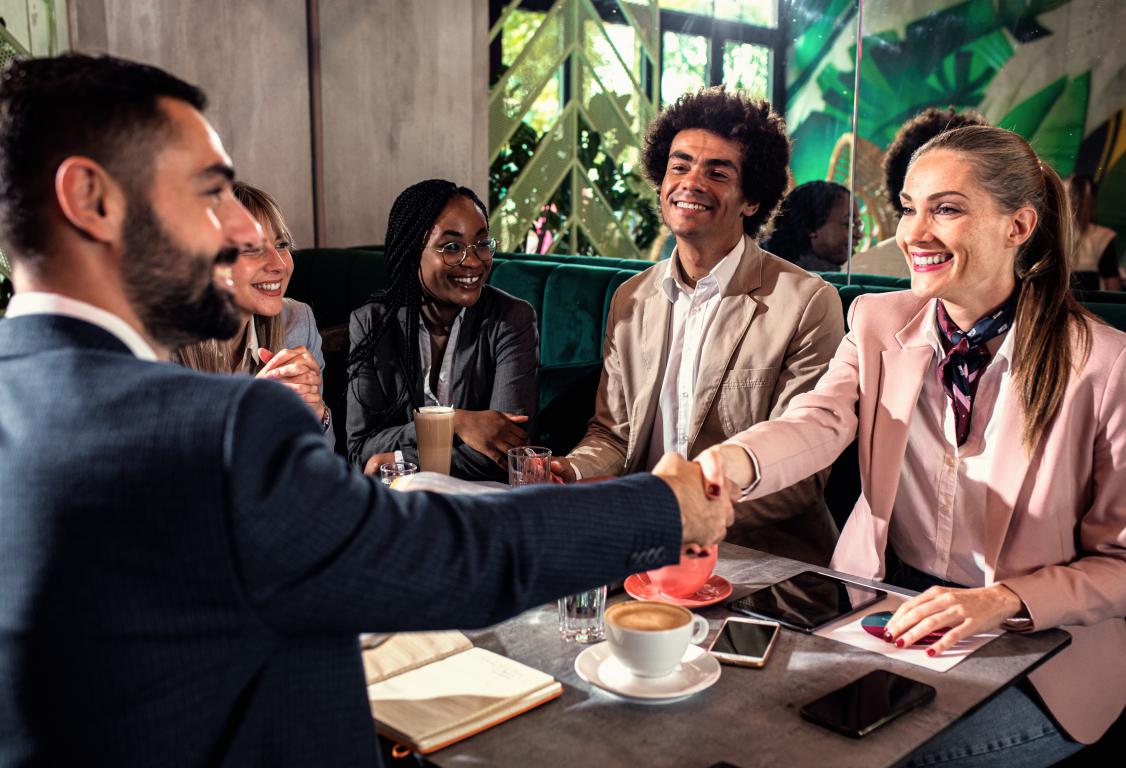 A diverse group of professionals smiles as a man and woman shake hands across a cafe table, finalizing a business agreement.