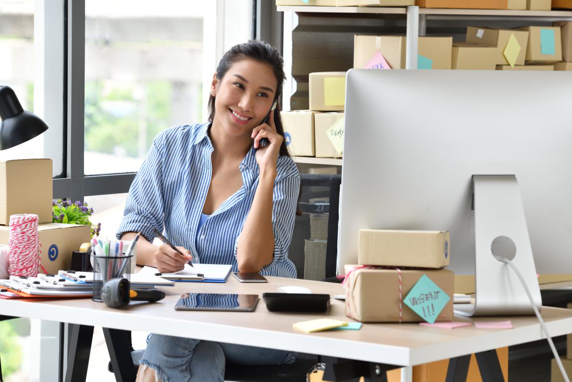 A smiling woman at a desk filled with shipping supplies talks on the phone while taking notes, looking happy and professional in her home office.