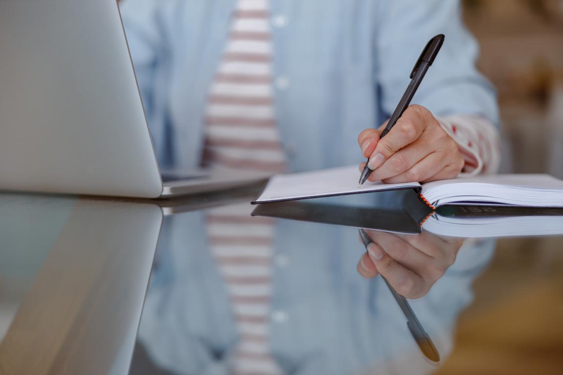 A close-up view of a person's hands writing notes in a physical notebook with a pen, positioned next to a laptop on a reflective desk.