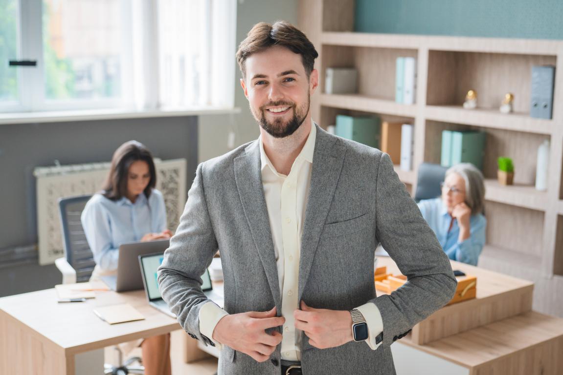 A confident young businessman in a grey blazer smiles warmly in a bright office, with colleagues working at their desks behind him.