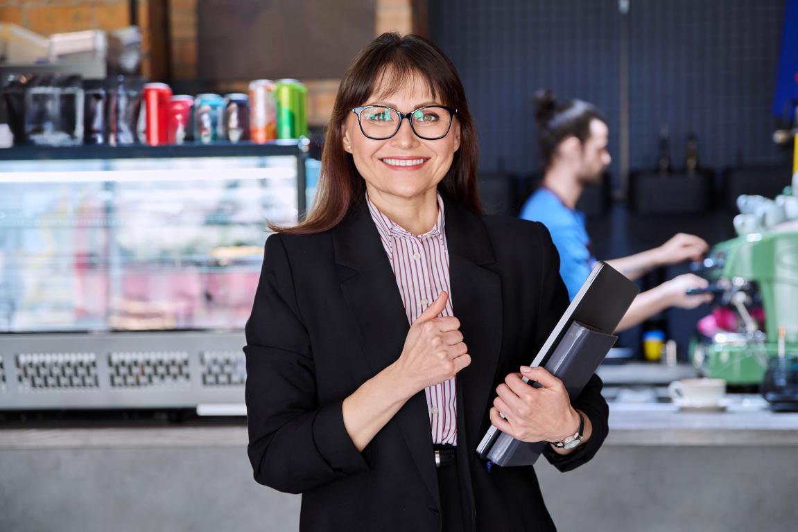 A smiling female manager in a café gives a thumbs-up while holding notebooks, projecting a sense of satisfaction and control with a barista working in the background.