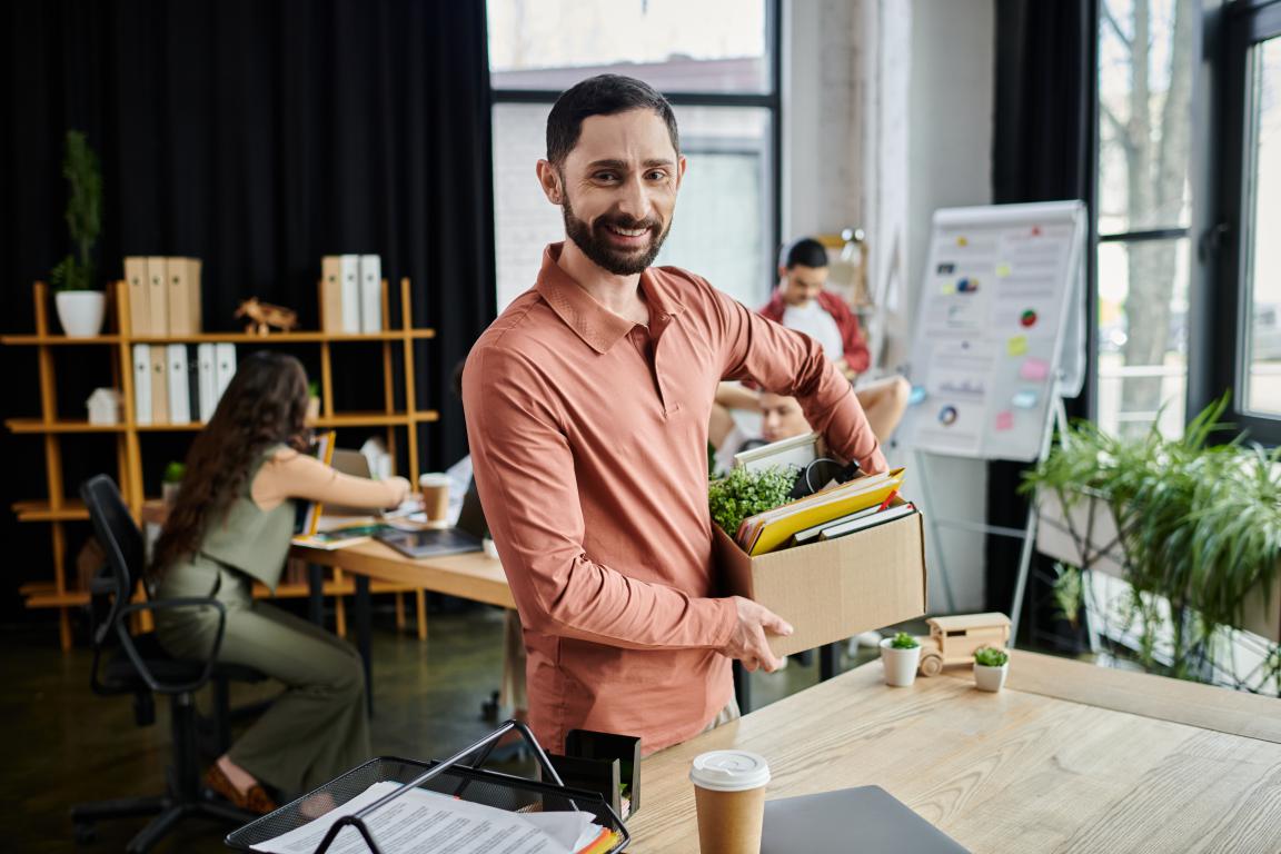A smiling man in a modern office holds a box of personal items, including files and a plant, suggesting a new beginning at work while colleagues are blurred in the background.