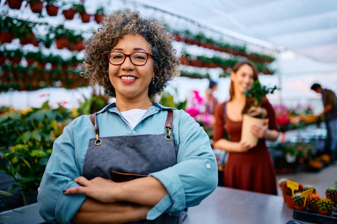 A friendly female business owner with curly hair and glasses smiles confidently in her garden centre, standing with her arms crossed in front of rows of plants.