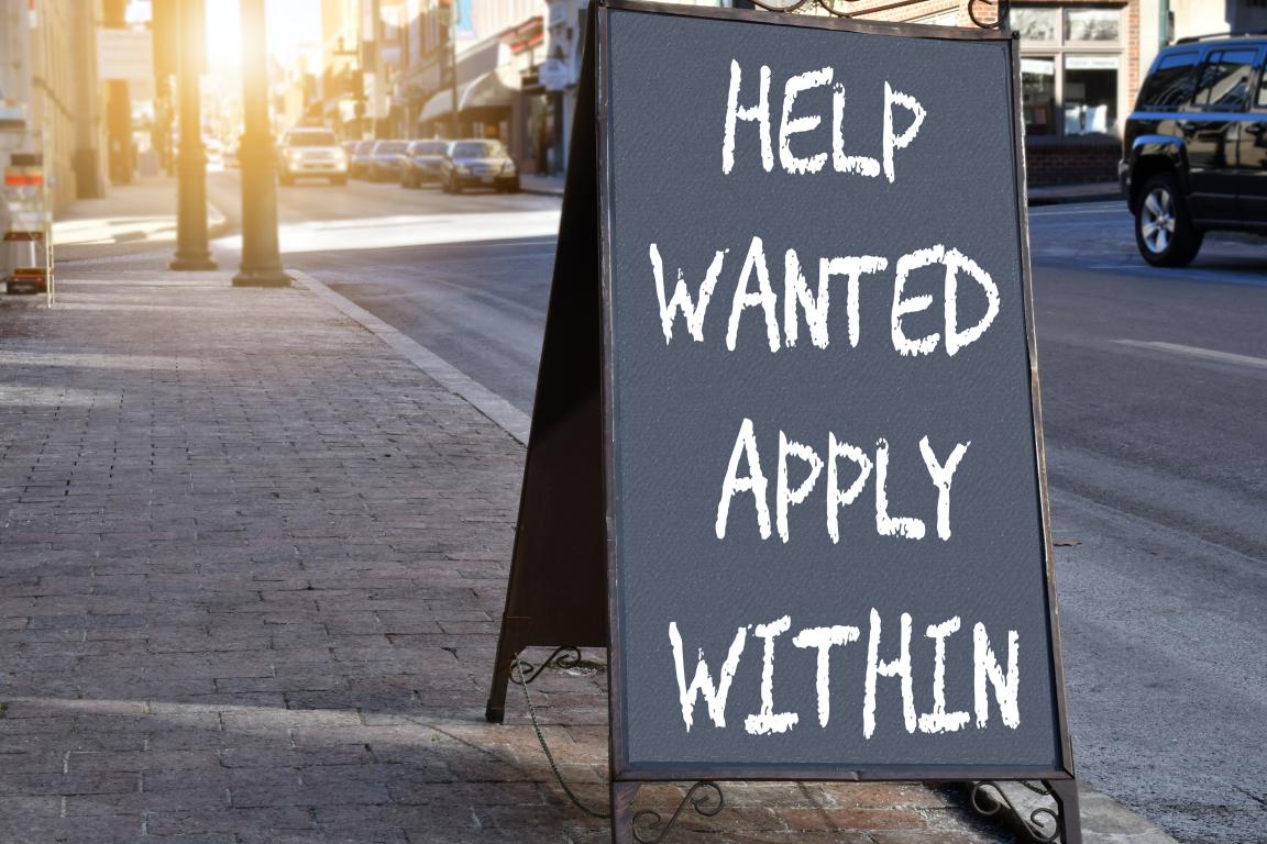 A black A-frame sign on a city street with the words "Help Wanted Apply Within" written in white.