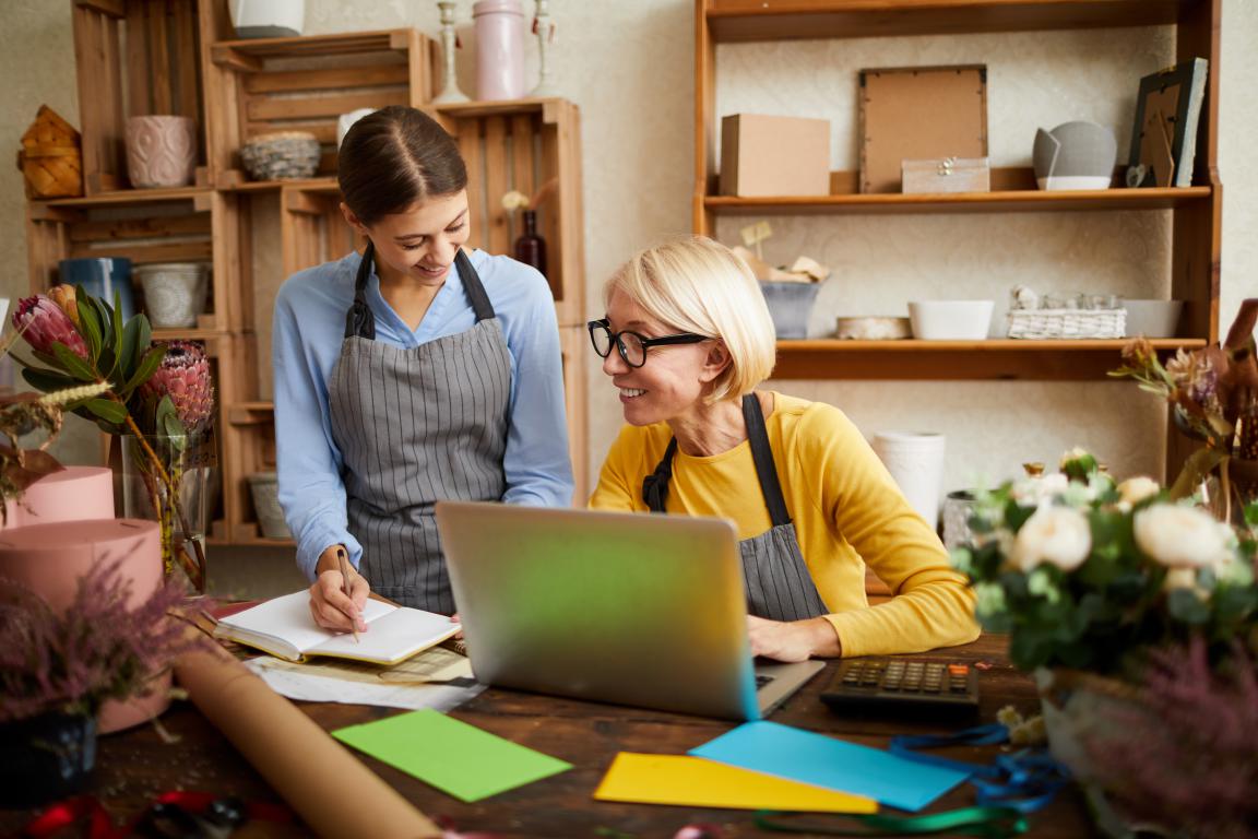  An experienced business owner trains a new employee in a craft workshop, smiling as the new hire takes notes.